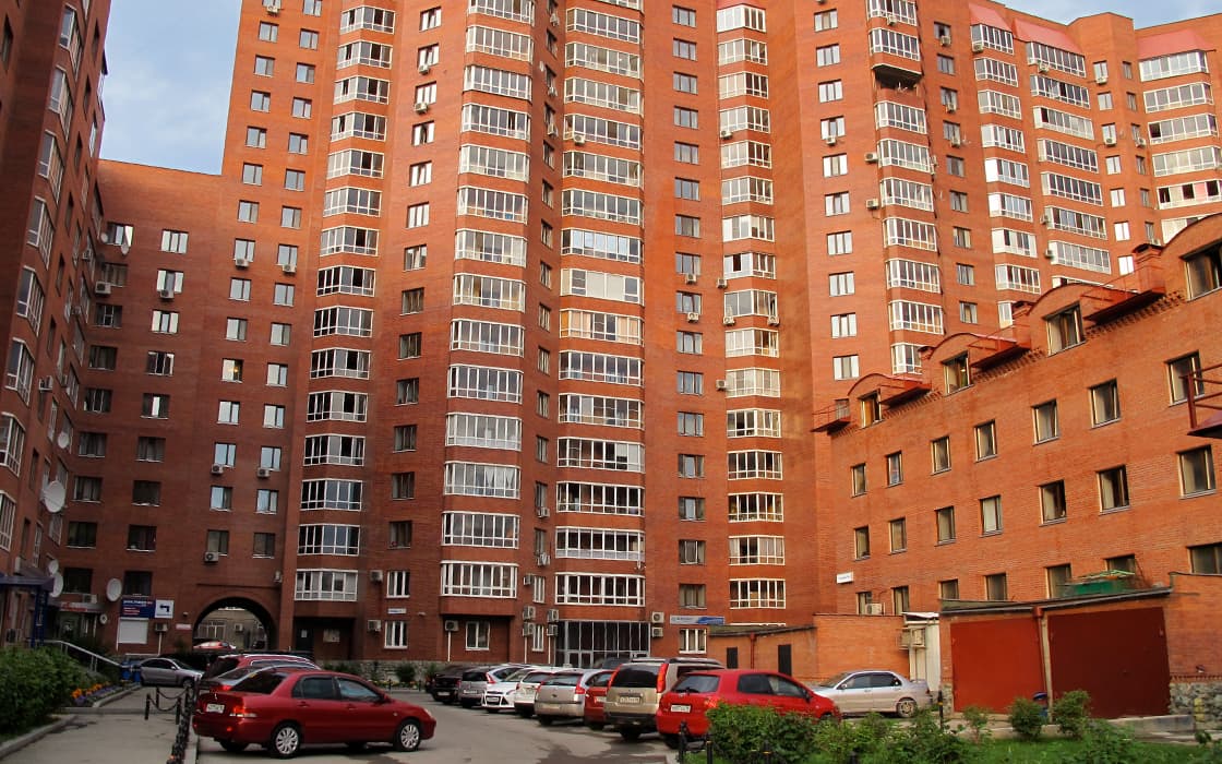 Hohe Wohnhäuser aus rotem Backstein mit Balkonen umgeben einen kleinen Innenhof mit geparkten Autos und Grünflächen.