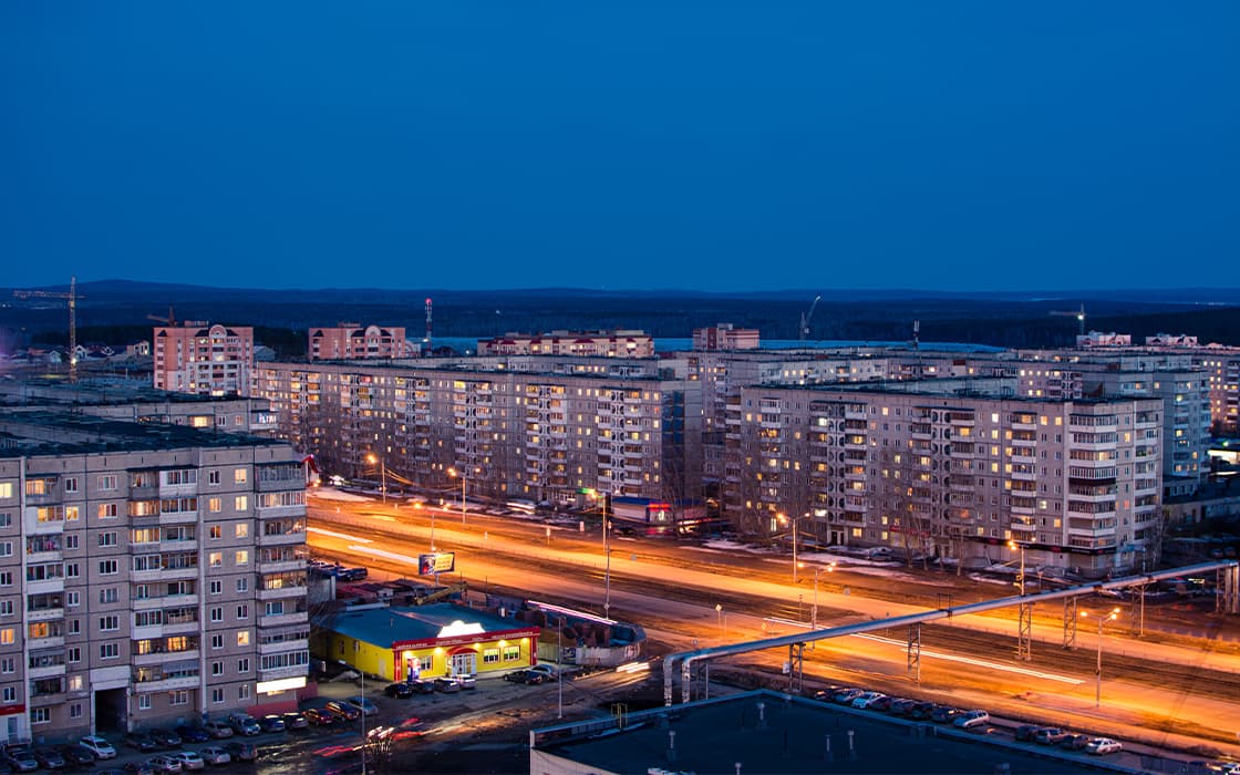 Ein Stadtbild in der Abenddämmerung mit Wohnhäusern und Lichtspuren von Autos auf einer belebten Straße.