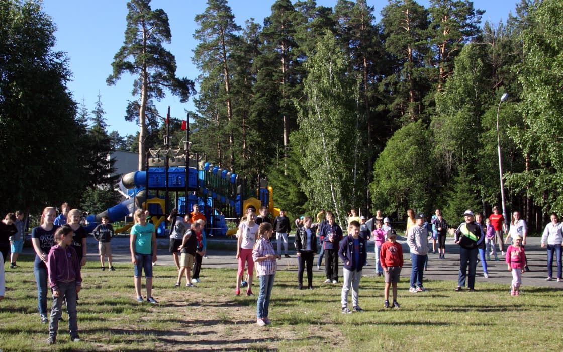 Kinder und Erwachsene stehen an einem sonnigen Tag mit Abstand in einem Park mit einem Spielplatz und Bäumen im Hintergrund.