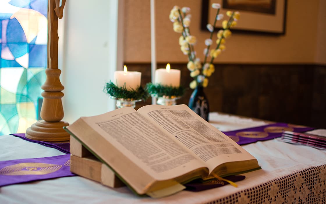 Eine aufgeschlagene Bibel auf einem Ständer, mit Kerzen, Blumen und einem Buntglasfenster im Hintergrund auf einem geschmückten Tisch.