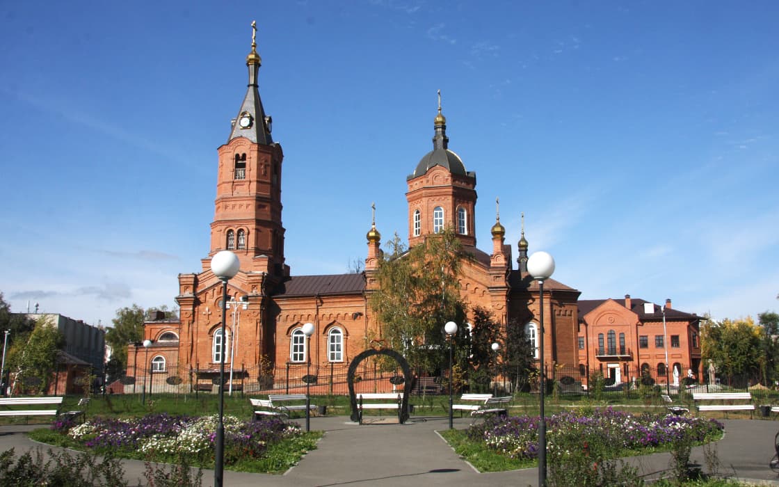 Kirche aus rotem Backstein mit zwei Türmen, Kuppeln und goldenen Kreuzen, umgeben von Bänken, Blumen und Laternenpfählen.