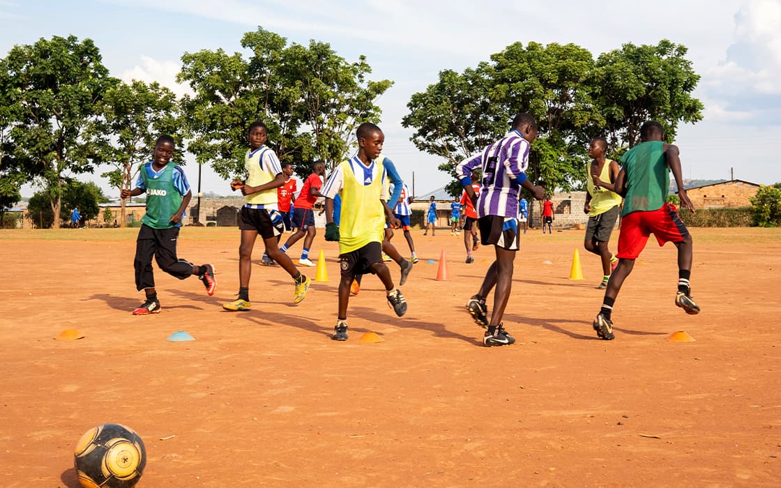 Kinder in Sportwesten spielen Fußball auf einem unbefestigten Feld mit Kegeln und Bäumen im Hintergrund.