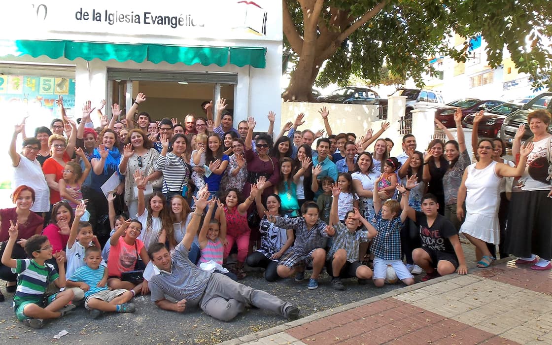 Eine große, bunt gemischte Gruppe von Menschen, die an einem sonnigen Tag vor einem Kirchengebäude lächeln und winken.