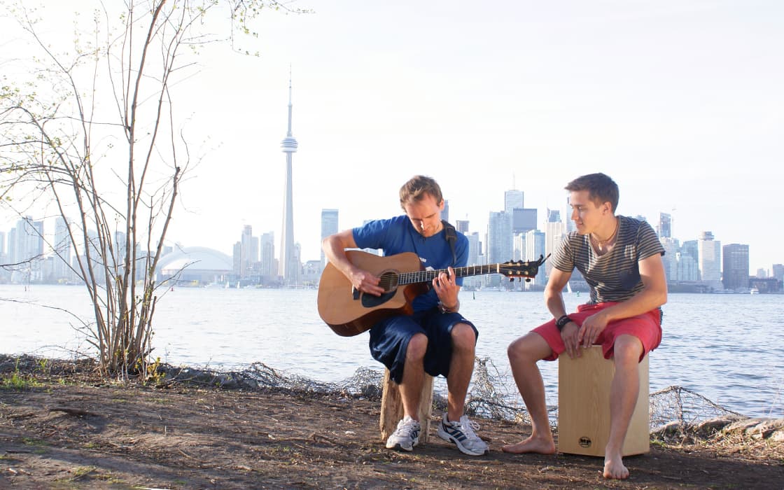 Zwei Männer spielen am Wasser mit Gitarre und Trommel Musik, im Hintergrund sind die Skyline von Toronto und der CN Tower zu sehen.