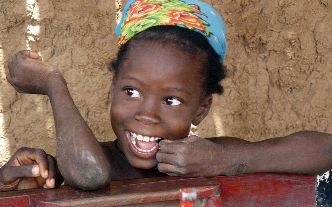 Ein lächelndes junges Mädchen mit einem bunten Kopftuch lehnt auf einer hölzernen Fläche neben einer strukturierten Lehmmauer.