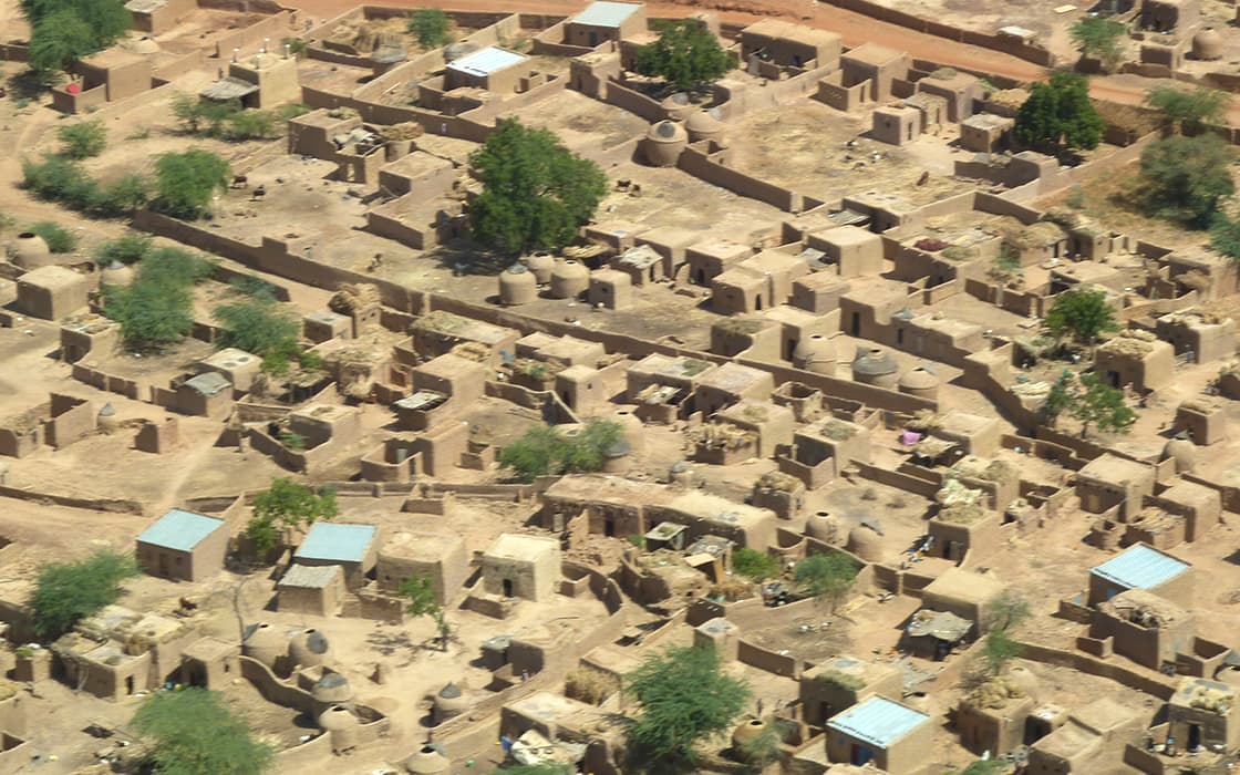 Luftaufnahme eines trockenen Dorfes mit Lehmziegelgebäuden, vereinzelten Bäumen und unbefestigten Wegen.