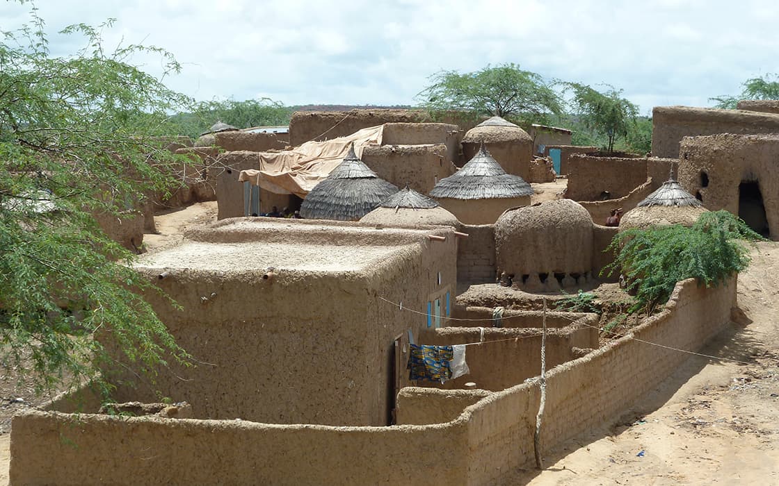 Lehmziegel- und Strohdachhäuser in einem ländlichen Dorf, umgeben von Grün unter einem bewölkten Himmel.