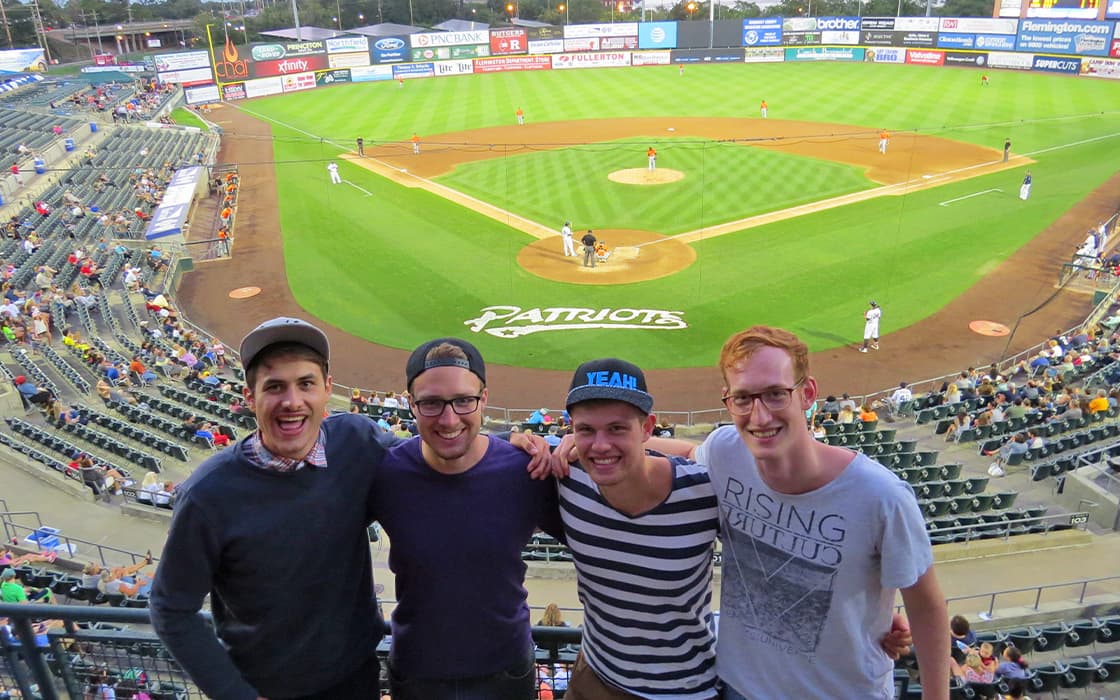 Vier lächelnde junge Männer posieren gemeinsam vor einem Baseballfeld während eines Spiels in einem Stadion.