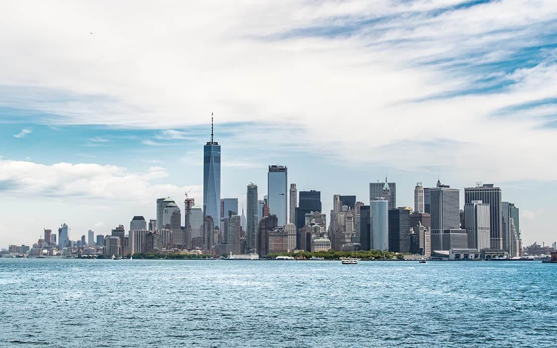 Die Skyline von New York City mit hohen Gebäuden und dem One World Trade Center über dem Wasser bei teilweise bewölktem Himmel.