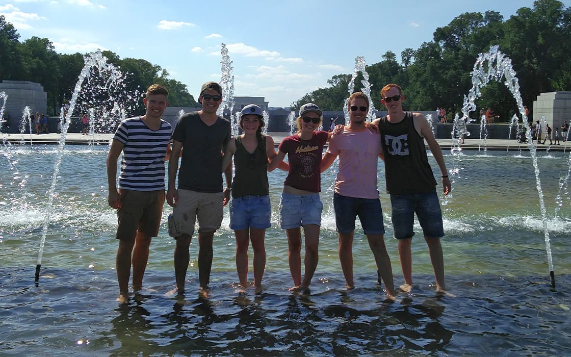 Sechs junge Menschen stehen an einem sonnigen Tag lächelnd in einem flachen Springbrunnen mit Wasserdüsen.