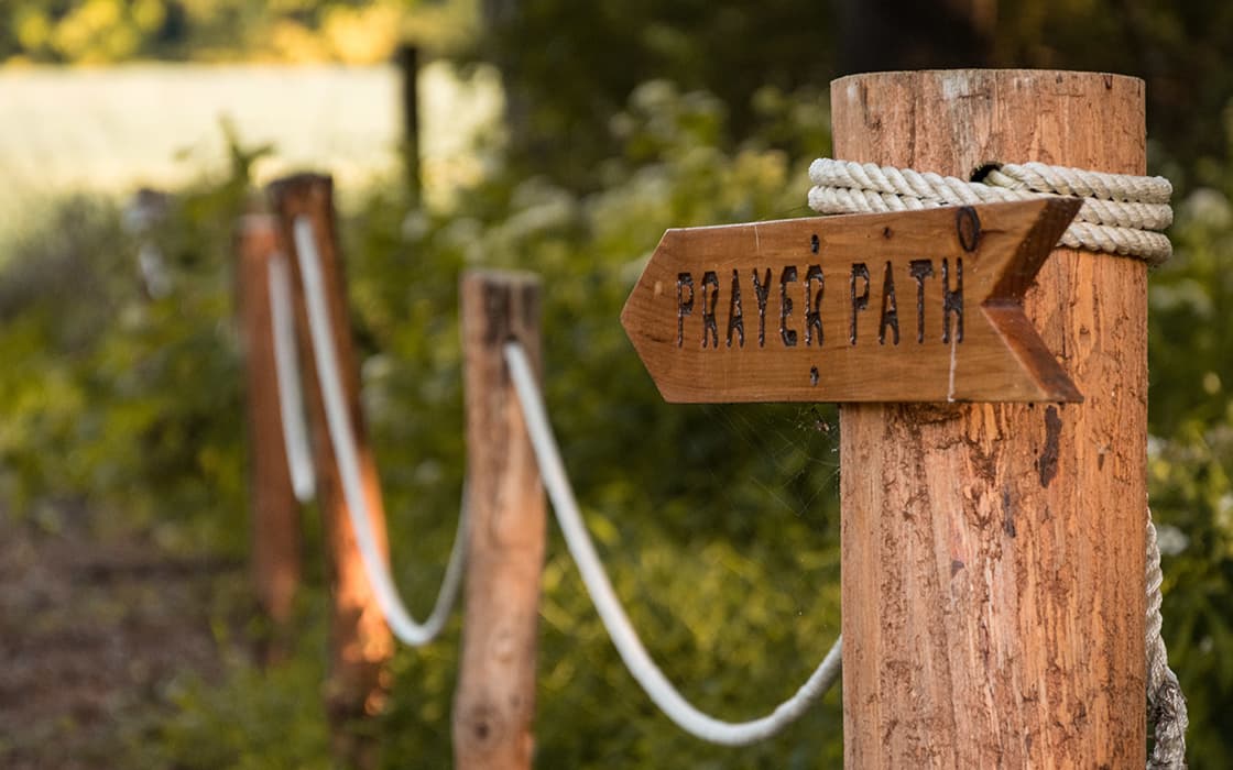 Ein hölzernes Schild mit der Aufschrift "Prayer Path" (Gebetsweg) weist auf einen mit Seilen gesäumten Pfad im Grünen hin.