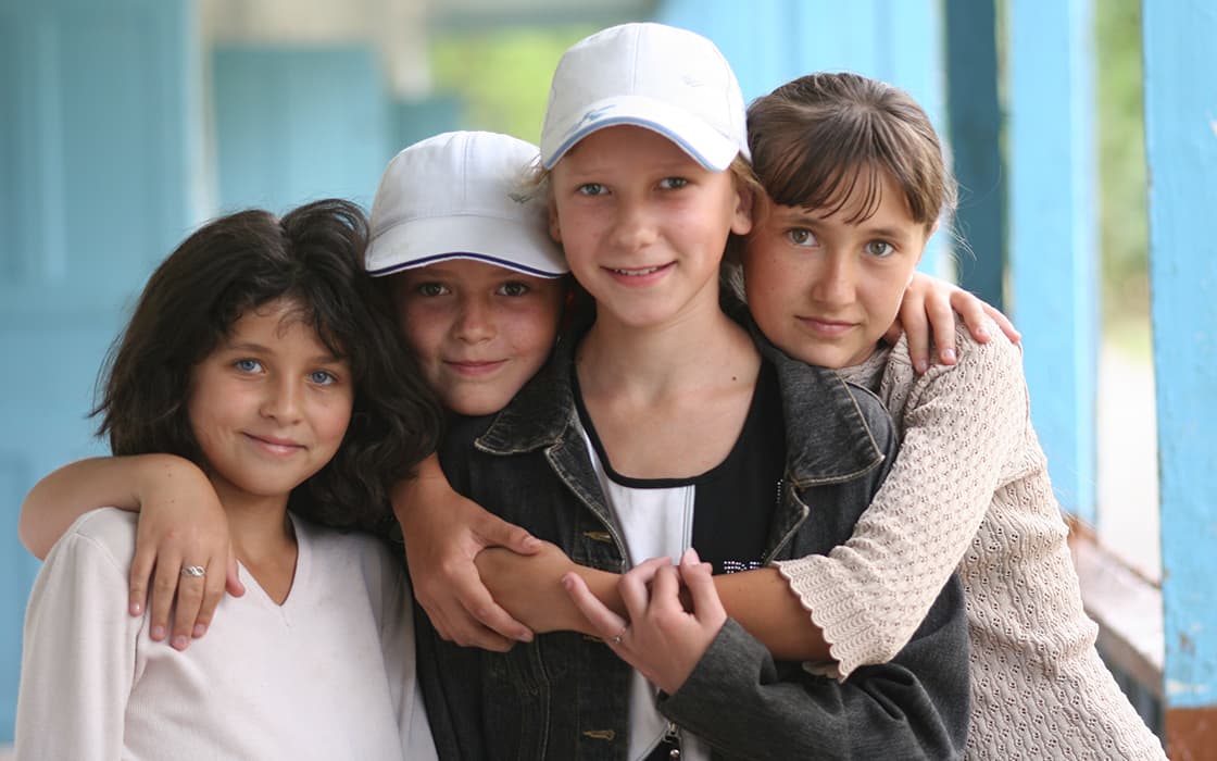 Vier Mädchen stehen dicht beieinander und lächeln in die Kamera, zwei von ihnen tragen weiße Baseballmützen.