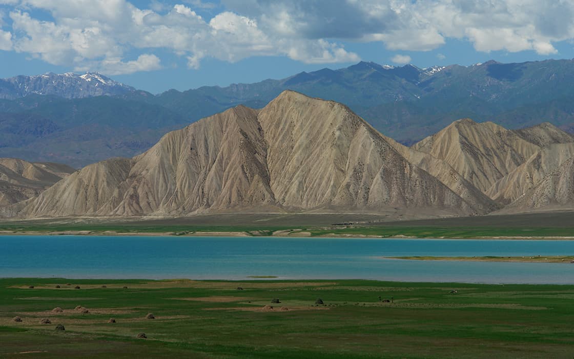Ein gestreifter Berg erhebt sich hinter einem blauen See und grünem Gras unter einem teilweise bewölkten Himmel mit entfernten Bergen.
