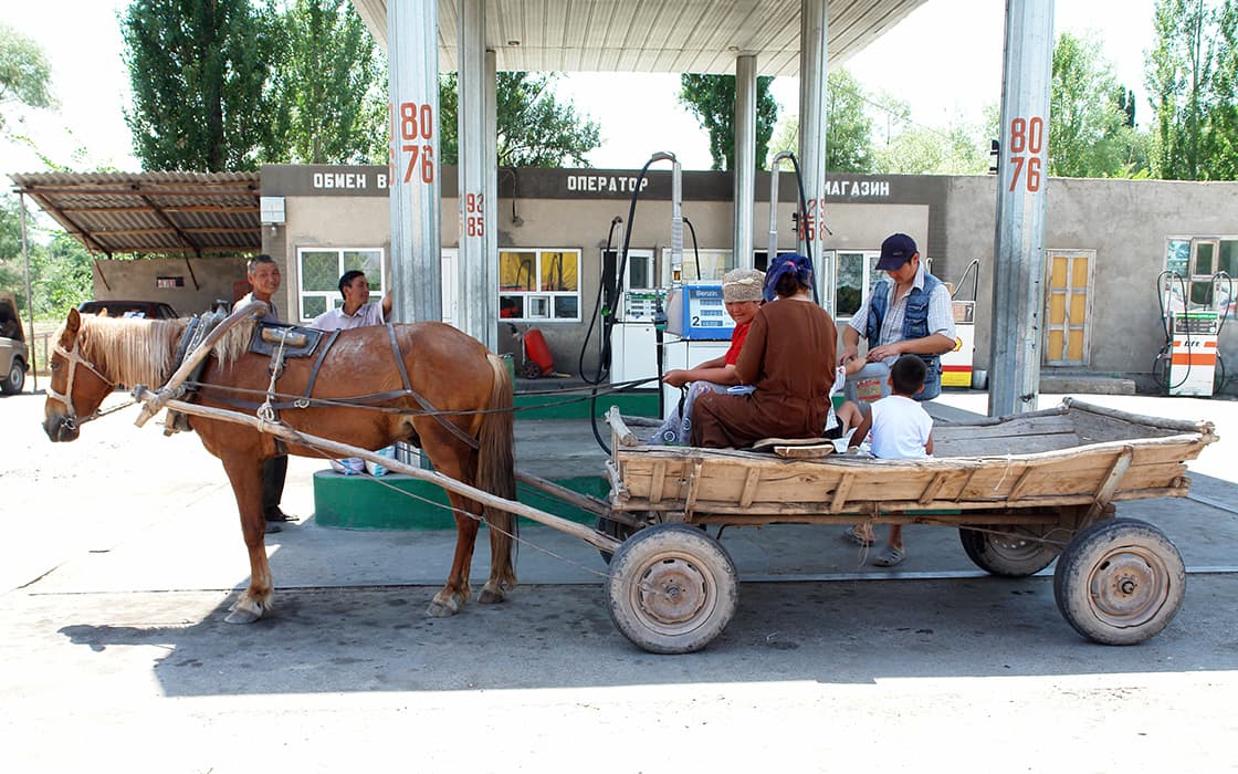 Ein hölzerner Pferdewagen mit Personen ist an einer Tankstelle mit Zapfsäulen und einem Gebäude im Hintergrund abgestellt.
