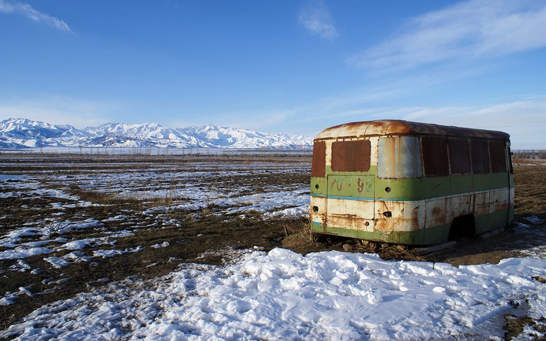 Ein alter, rostiger, grüner Bus steht auf einem verschneiten Feld mit Bergen im Hintergrund unter blauem Himmel.