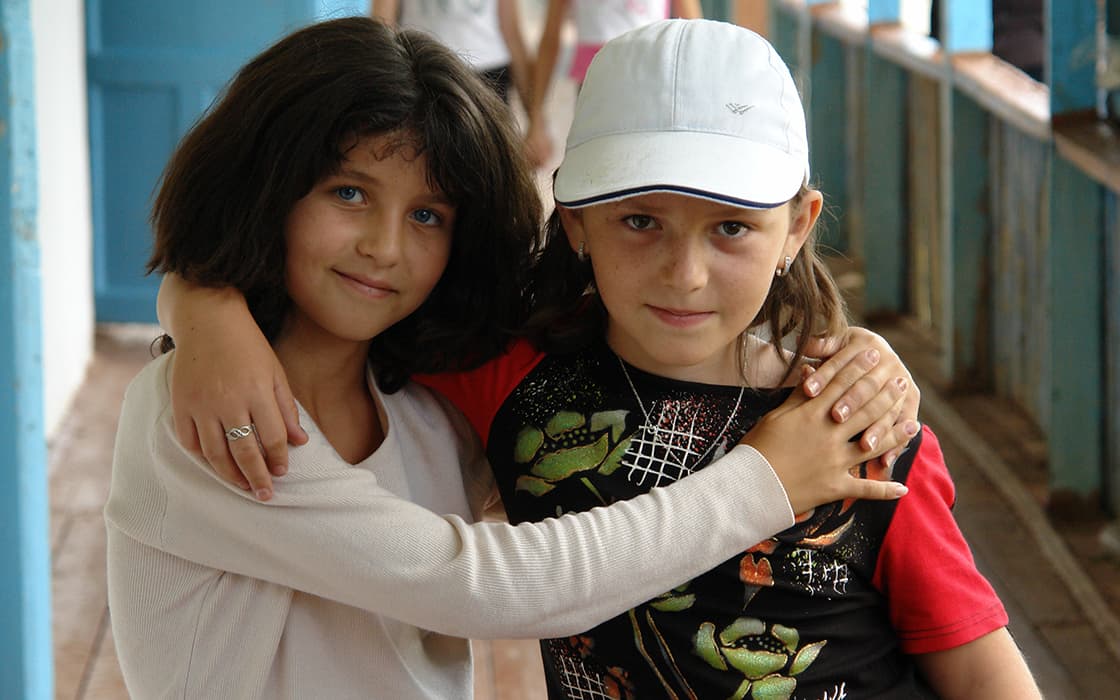 Zwei Mädchen stehen eng beieinander, die Arme umeinander gelegt, und lächeln in die Kamera in einem Hausflur.