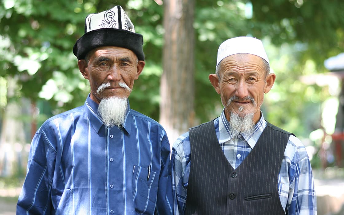 Zwei ältere Männer mit langen weißen Bärten, die traditionelle Hüte und Hemden tragen, stehen im Freien mit Bäumen im Hintergrund.