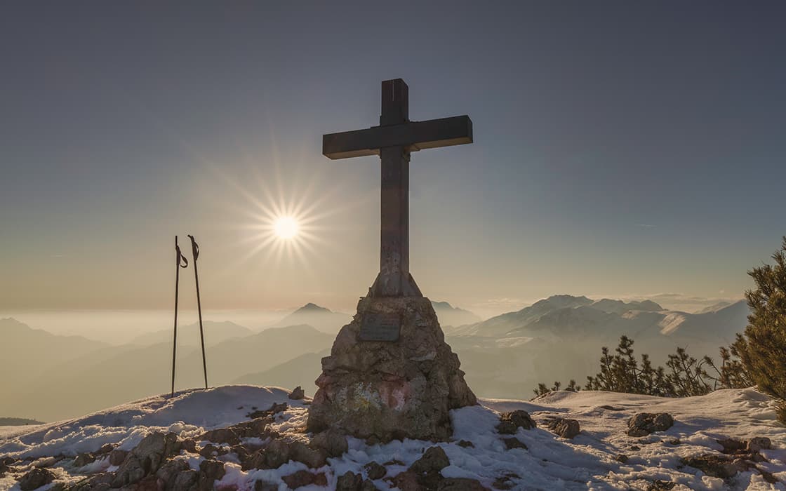Ein großes Kreuz auf einem verschneiten Berggipfel bei Sonnenaufgang, mit Skistöcken und fernen Bergen im Hintergrund.