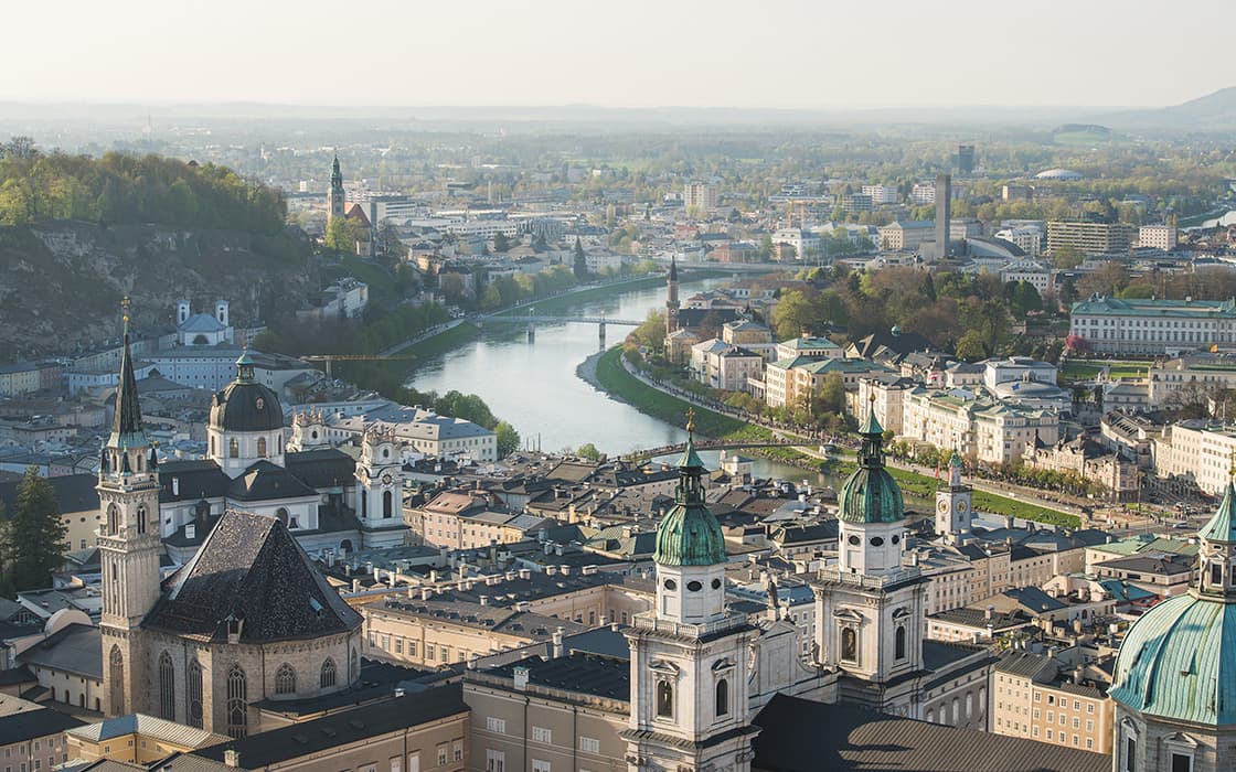 Luftaufnahme von Salzburg, Österreich, mit einem Fluss, historischen Gebäuden und entfernten Hügeln unter einem dunstigen Himmel.