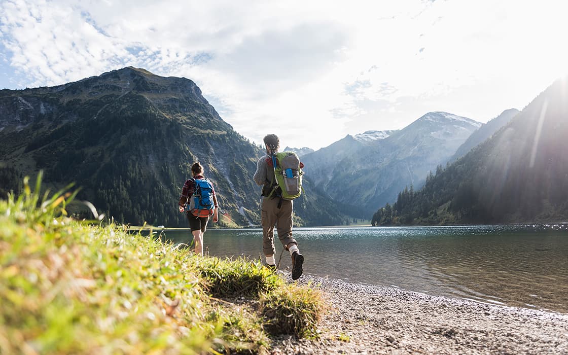 Zwei Wanderer mit Rucksäcken gehen auf einen Bergsee zu, umgeben von grasbewachsenen Hügeln und bewaldeten Bergen unter einem strahlenden Himmel.