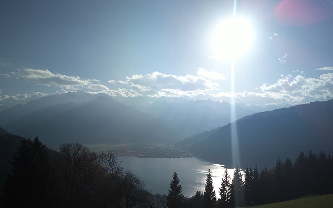 Die Sonne scheint über einem See, der von Bergen und Kiefern umgeben ist, mit vereinzelten Wolken am Himmel.