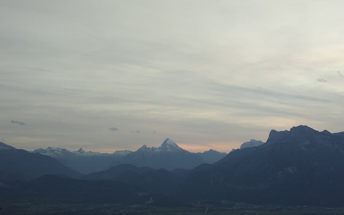 Entfernte schneebedeckte Berge unter einem bewölkten Himmel in der Abenddämmerung, mit sanften Hügeln und Tälern im Vordergrund.