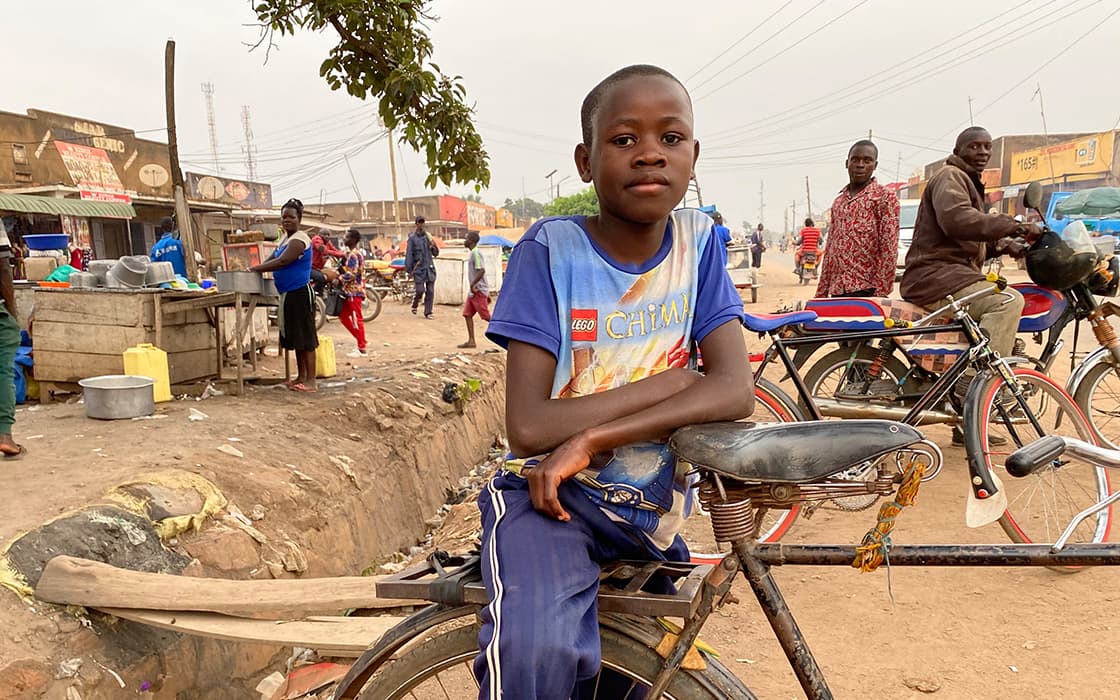 Ein Junge in einem blauen Hemd sitzt auf einem Fahrrad in einer belebten Straße mit Menschen, Geschäften und Fahrrädern im Hintergrund.