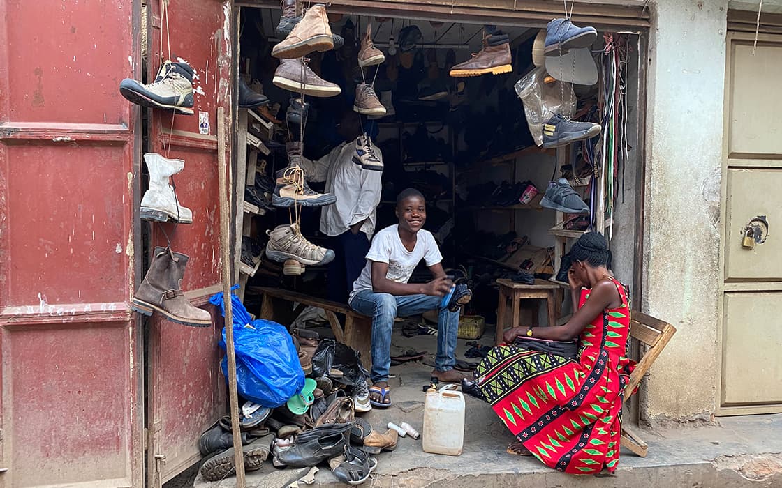 Ein Junge lächelt in einer Schuhreparaturwerkstatt, in der Schuhe herumstehen, während eine Frau in einem gemusterten Kleid in der Nähe sitzt.