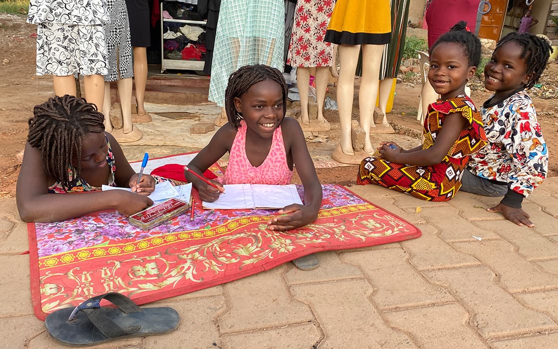 Vier junge Mädchen lächeln und lernen im Freien auf einer Matte vor einem Bekleidungsgeschäft mit Schaufensterpuppen im Hintergrund.