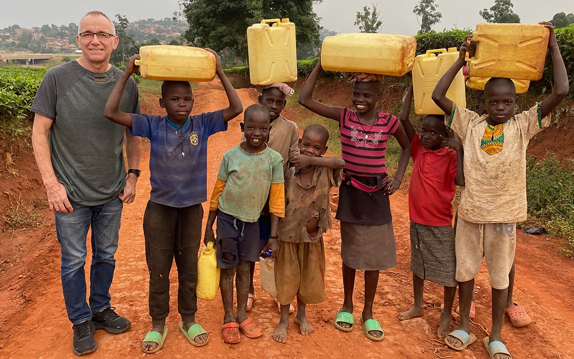 Ein Mann und fröhliche Kinder stehen zusammen auf einem Feldweg. Einige Kinder balancieren gelbe Wasserbehälter auf ihren Köpfen.