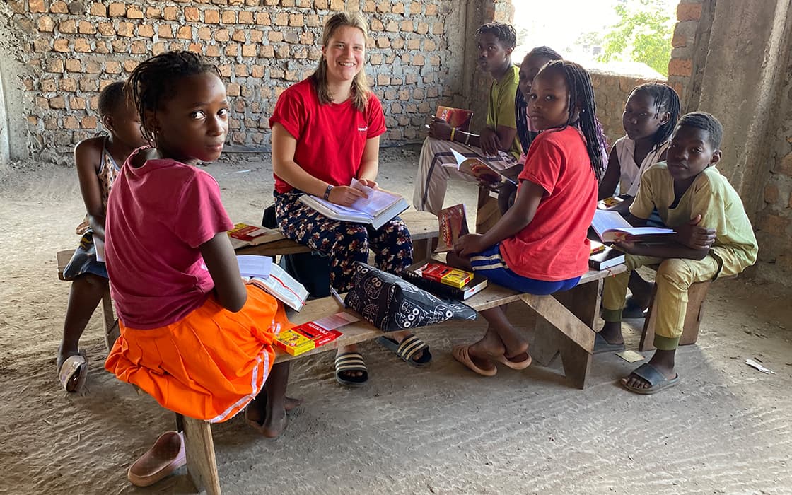 Eine Gruppe von Kindern und ein Erwachsener sitzen auf Bänken in einem rustikalen Klassenzimmer, schreiben in Hefte und schauen in die Kamera.