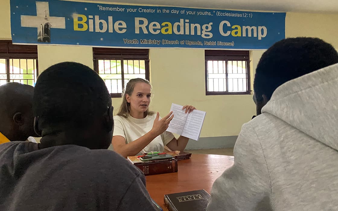 Eine Frau leitet eine Bibelstunde mit drei Jugendlichen an einem Tisch unter einem Bibellesecamp-Banner.