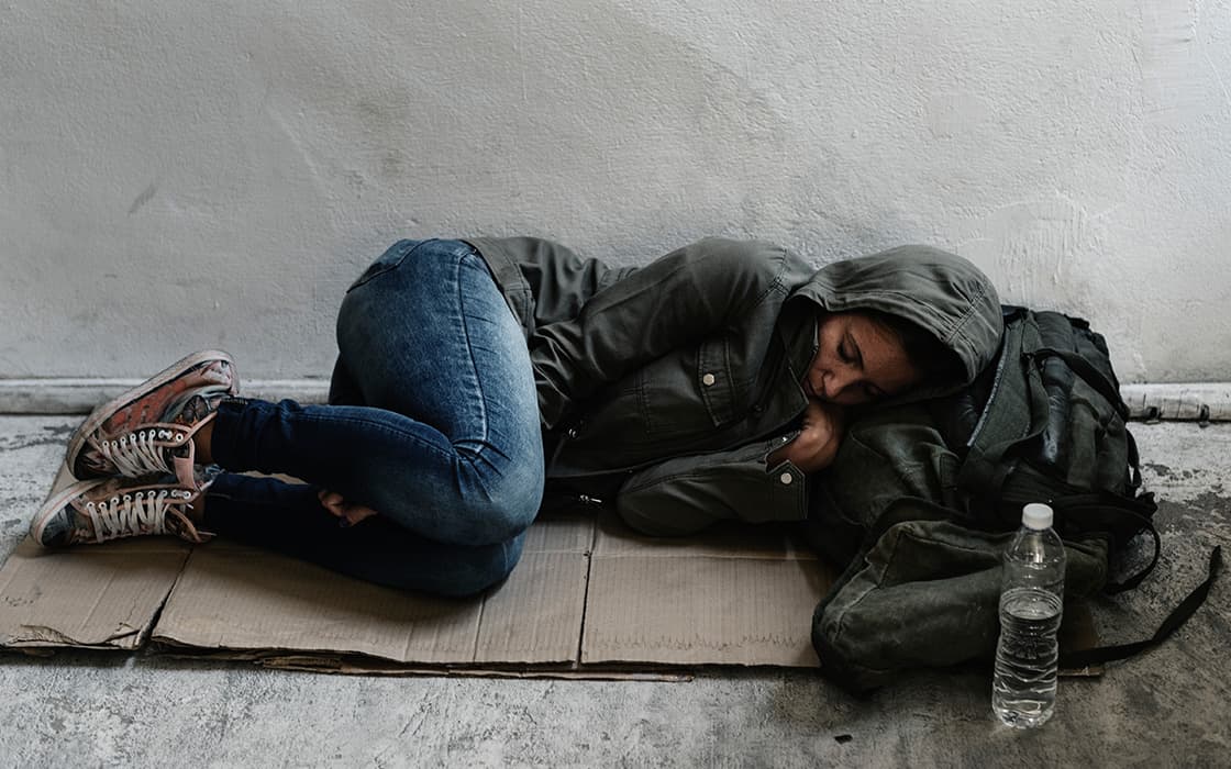 Eine Person im Kapuzenpulli schläft auf einem Karton in Griechenland, neben sich einen Rucksack und eine Wasserflasche.