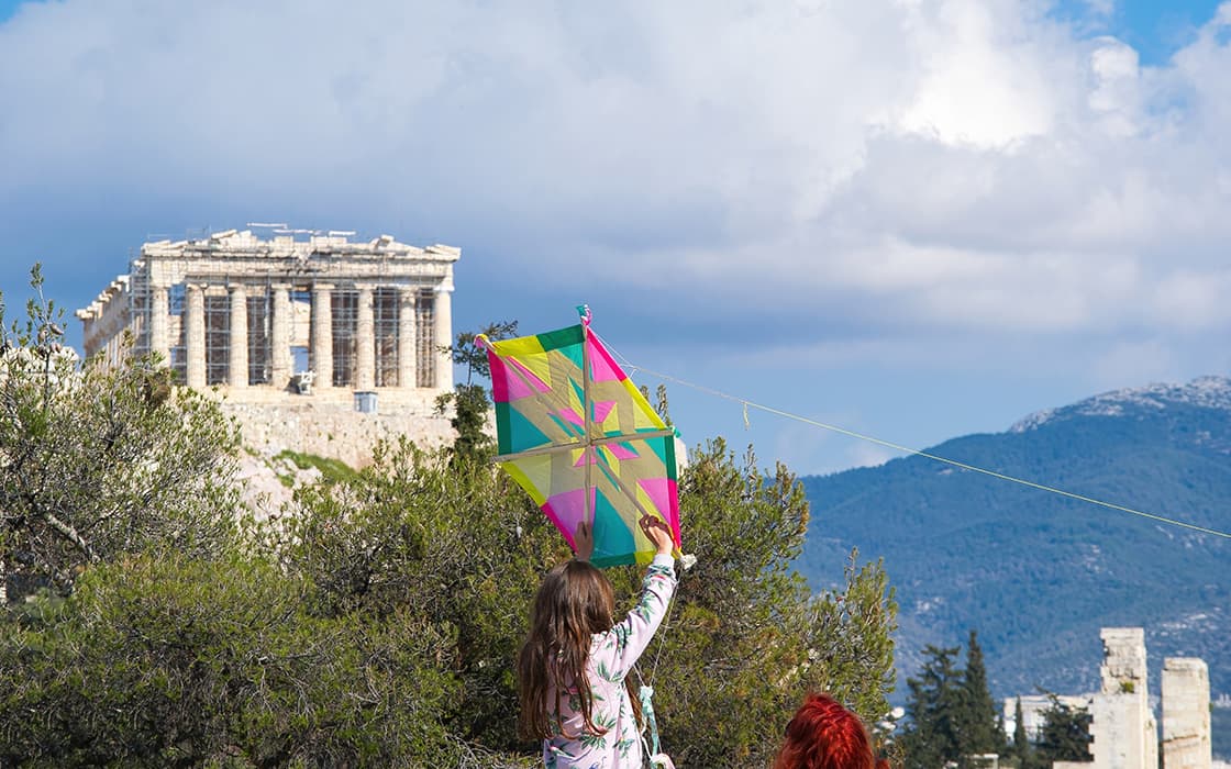 Ein Kind lässt einen bunten Drachen in der Nähe des Parthenon in Griechenland steigen, mit Bergen und blauem Himmel im Hintergrund.