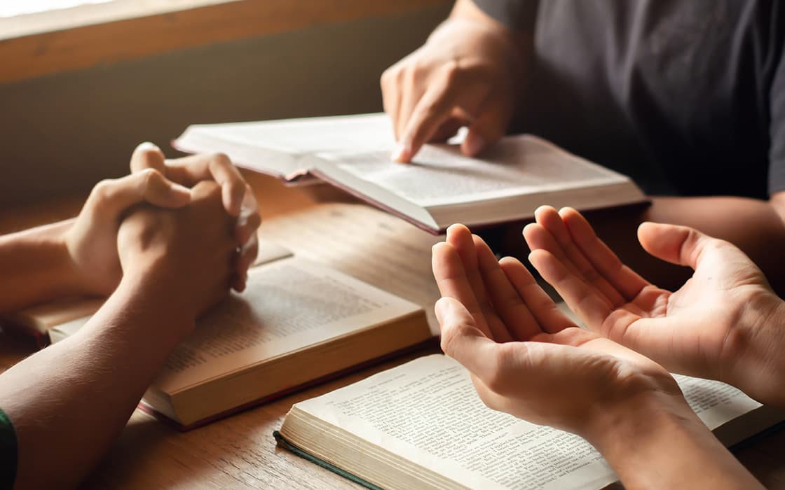 Drei Menschen in Griechenland sitzen an einem Tisch mit aufgeschlagenen Büchern und diskutieren, während der eine gestikuliert und der andere auf eine Seite zeigt.