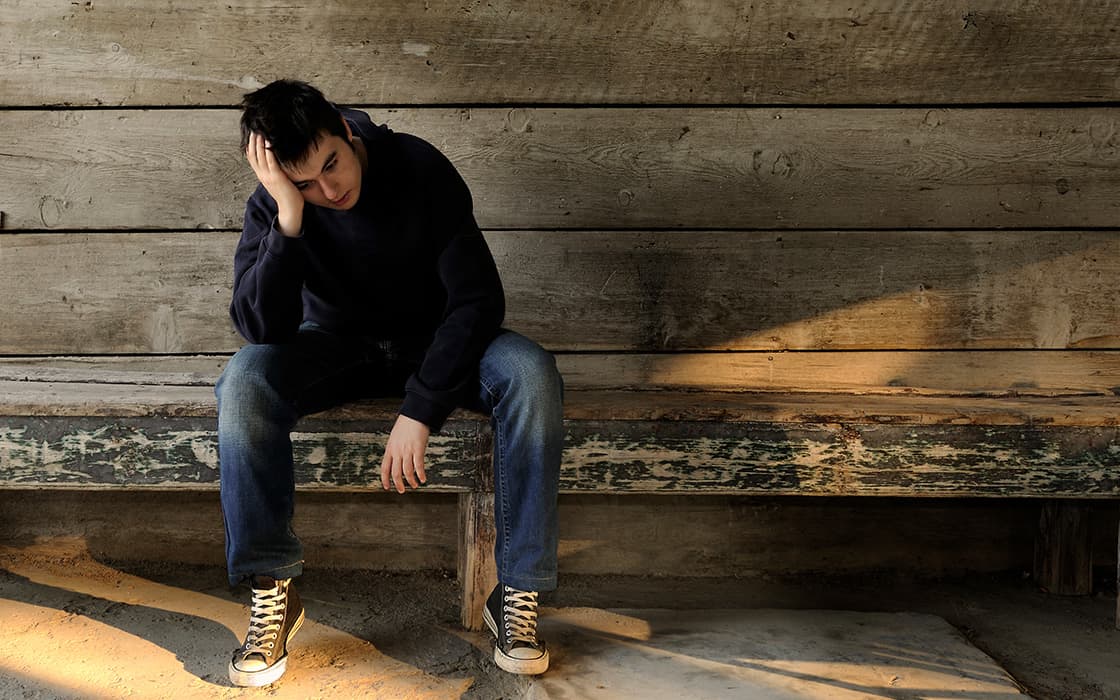 Eine Person sitzt allein auf einer Holzbank in Griechenland, den Kopf auf eine Hand gestützt, und scheint traurig oder in Gedanken versunken zu sein.