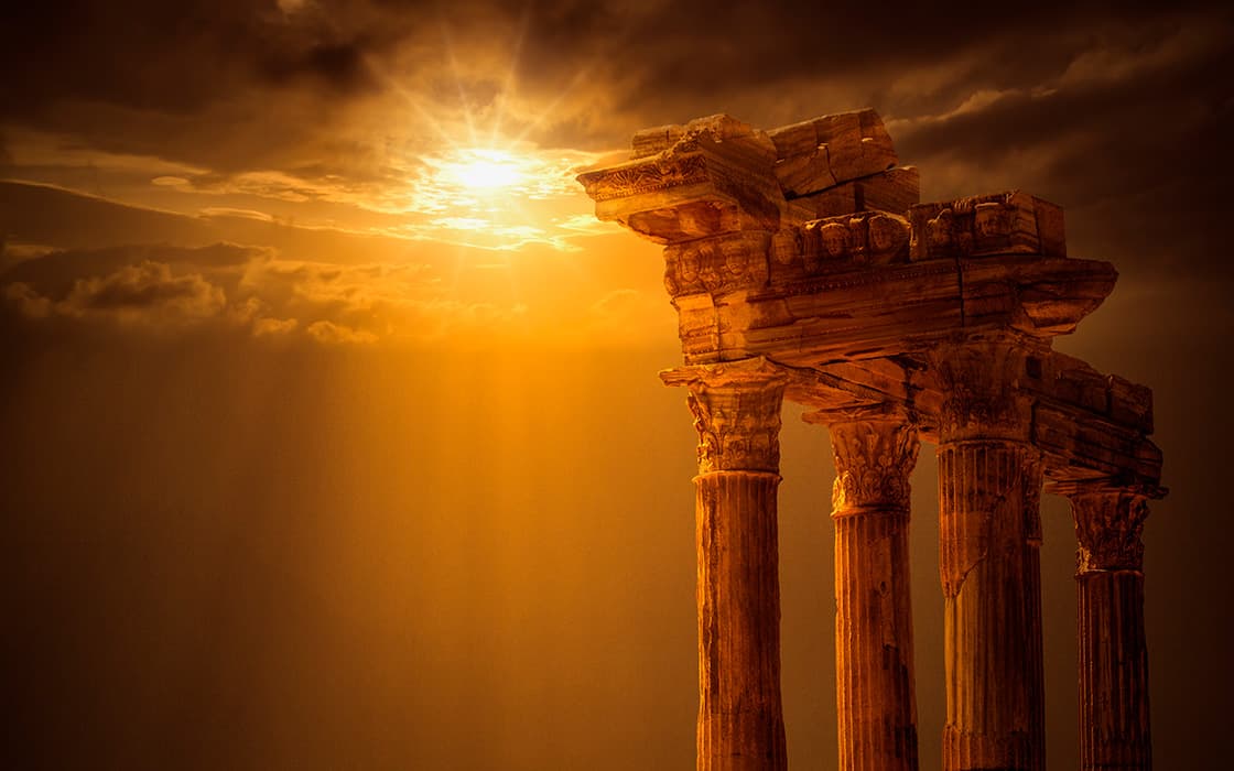 Antike griechische Tempelruinen in Griechenland mit drei Säulen bei Sonnenuntergang, goldenes Sonnenlicht bricht durch dramatische Wolken.