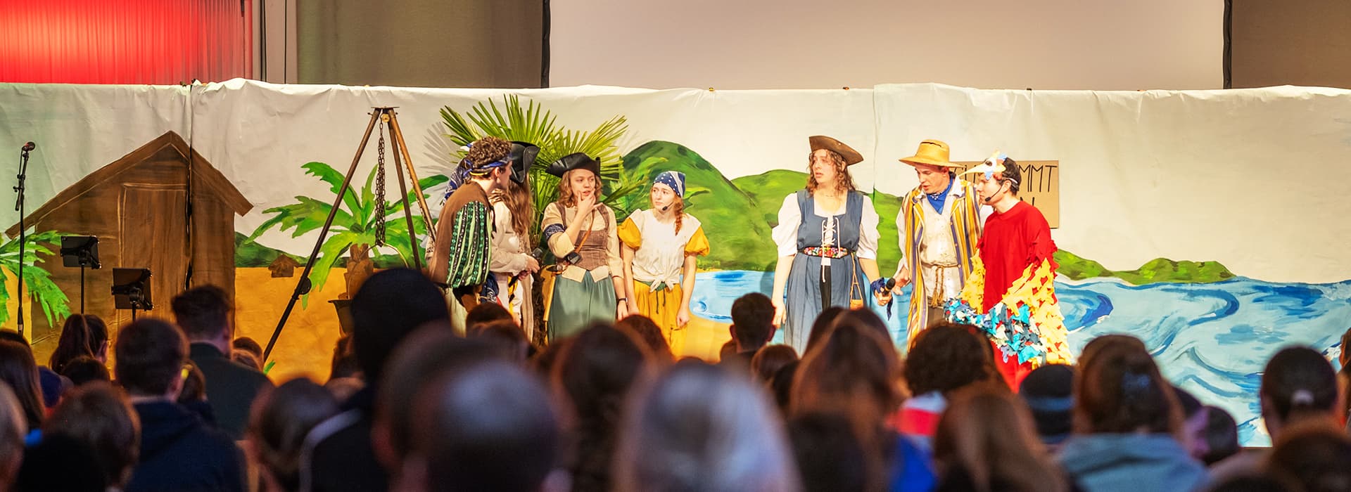 Schauspieler in farbenfrohen Piratenkostümen treten auf der Bühne vor einer gemalten tropischen Kulisse vor einem Publikum auf.