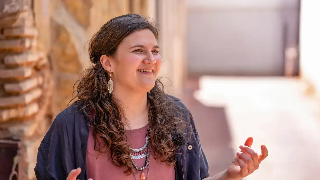 Rebekka Eggeler lächelt und gestikuliert mit ihren Händen im Freien in Spanien, wo sie an einer Steinmauer steht.