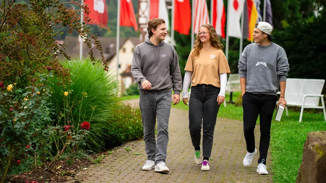 Drei junge Erwachsene lächeln auf einem Weg, der von Grünpflanzen, internationalen Flaggen gesäumt ist.