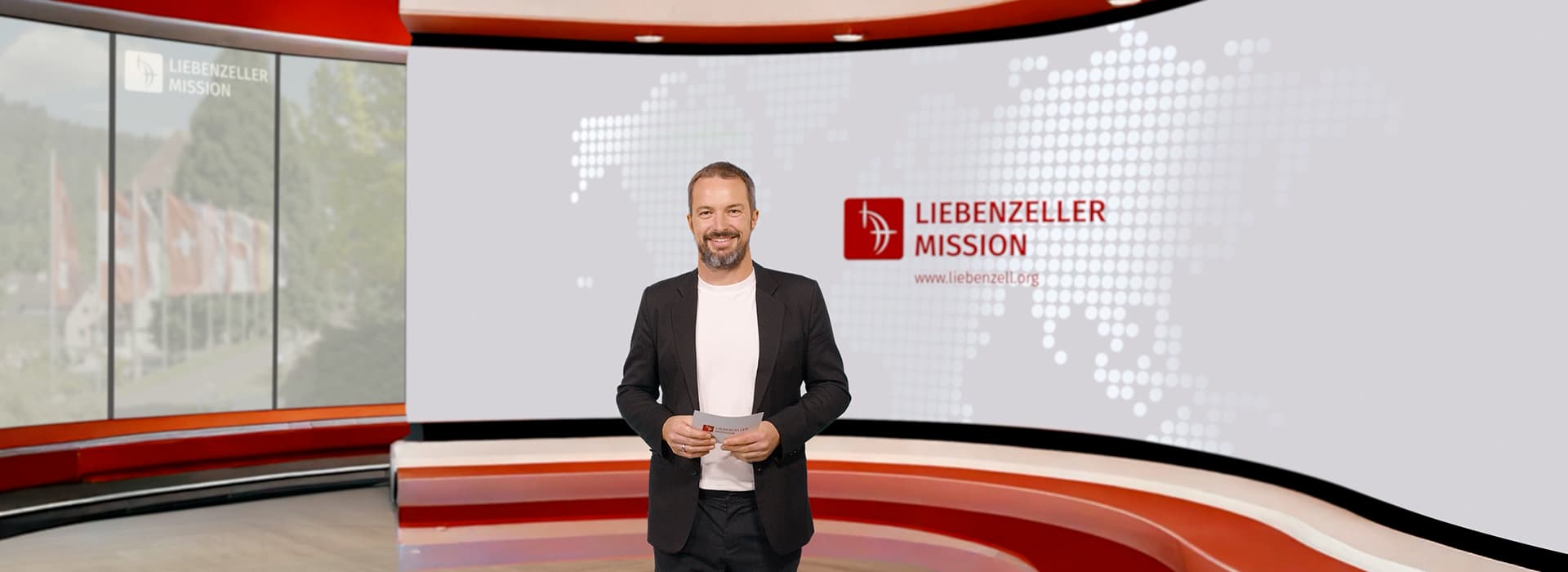 Ein Mann im Anzug steht in einem modernen Studio, hinter ihm das Logo der Liebenzeller Mission und eine Weltkarte auf dem Bildschirm.