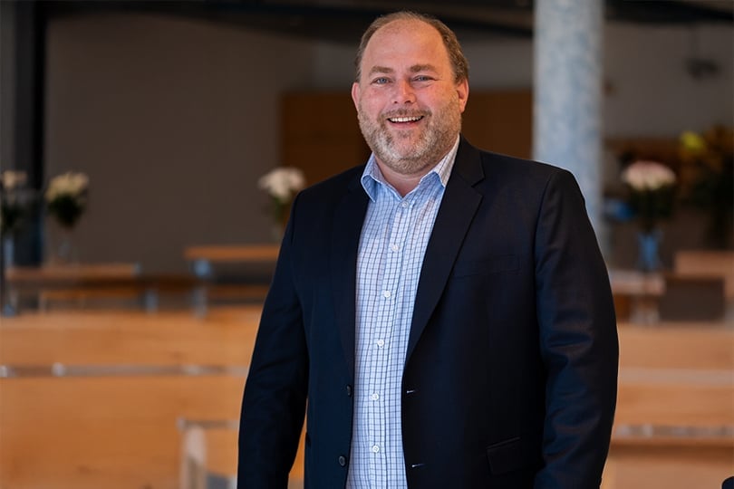 Ein lächelnder Mann in Anzugjacke und kariertem Hemd steht in dem modernen, vom Himmel beleuchteten Innenraum des MODAL 2025.