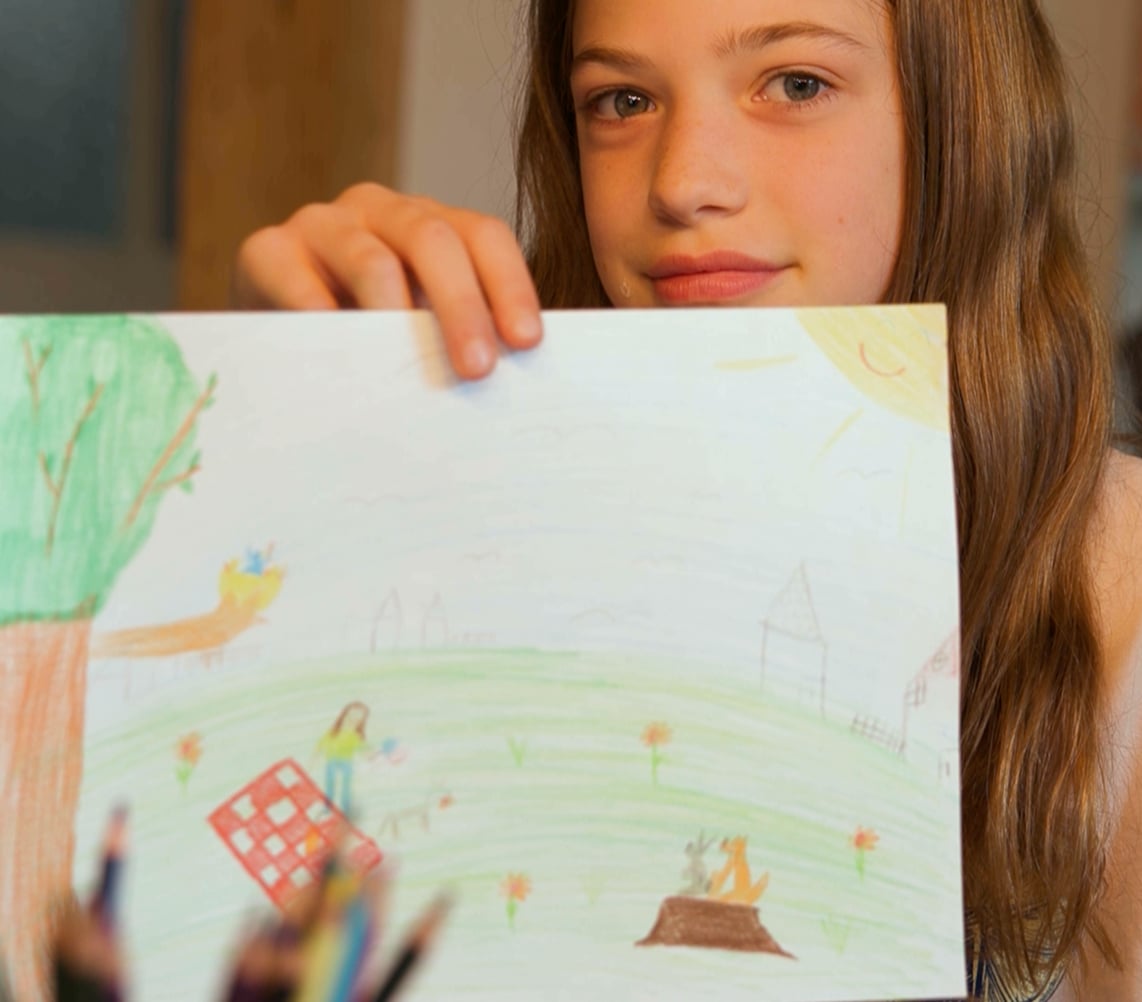 Ein Mädchen hält eine bunte Zeichnung von einem Baum, Menschen und Tieren unter dem Himmel hoch.