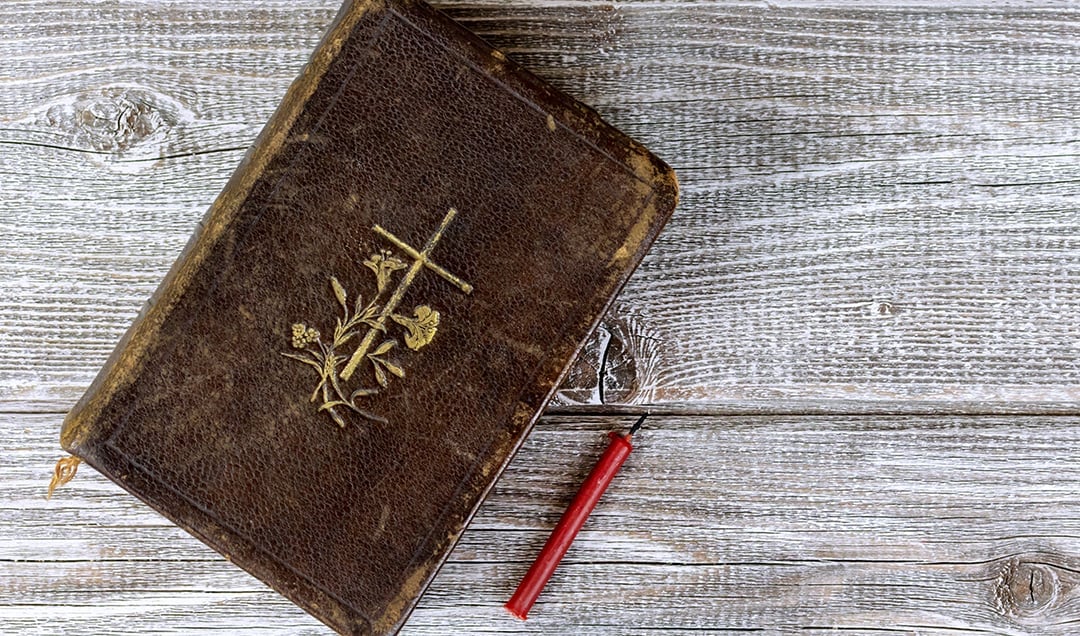 Eine alte braune Lederbibel mit einem goldenen Kreuz und einer Blume, daneben ein roter Stift auf Holz.