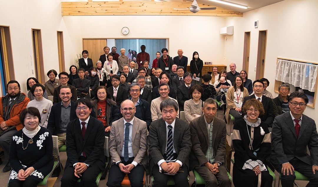 Eine förmlich gekleidete Gruppe sitzt und steht in Reihen in einem gut beleuchteten Raum und lächelt in die Kamera.