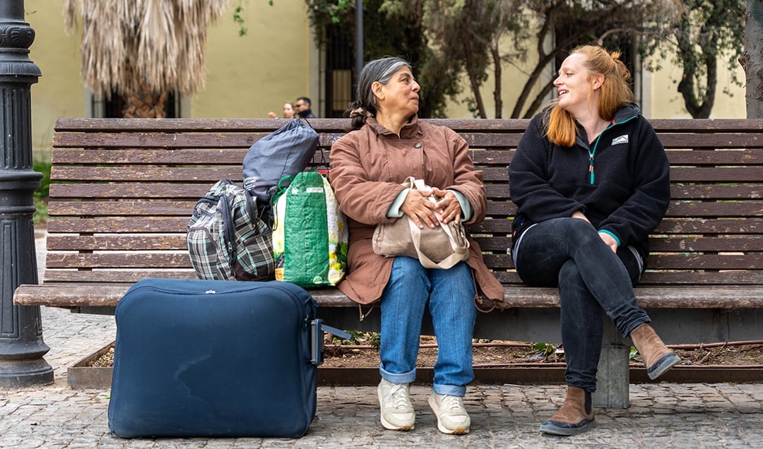 Zwei Frauen sitzen auf einer Bank mit Gepäck, lächeln und unterhalten sich im Freien in der Nähe des Geländes.