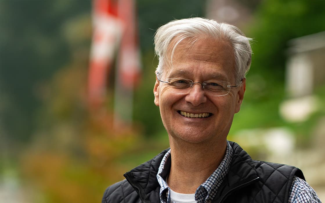 Lächelnder älterer Mann mit Brille und grauem Haar im Freien, mit Grünpflanzen und einer verschwommenen rot-weißen Flagge im Hintergrund.
