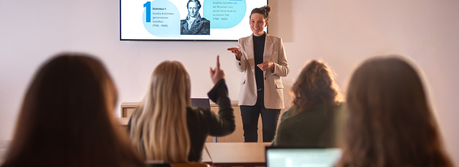 Ein Lehrer hält einen Vortrag, während die Schüler zuhören, und ein Schüler hebt die Hand, um eine Frage zu stellen.