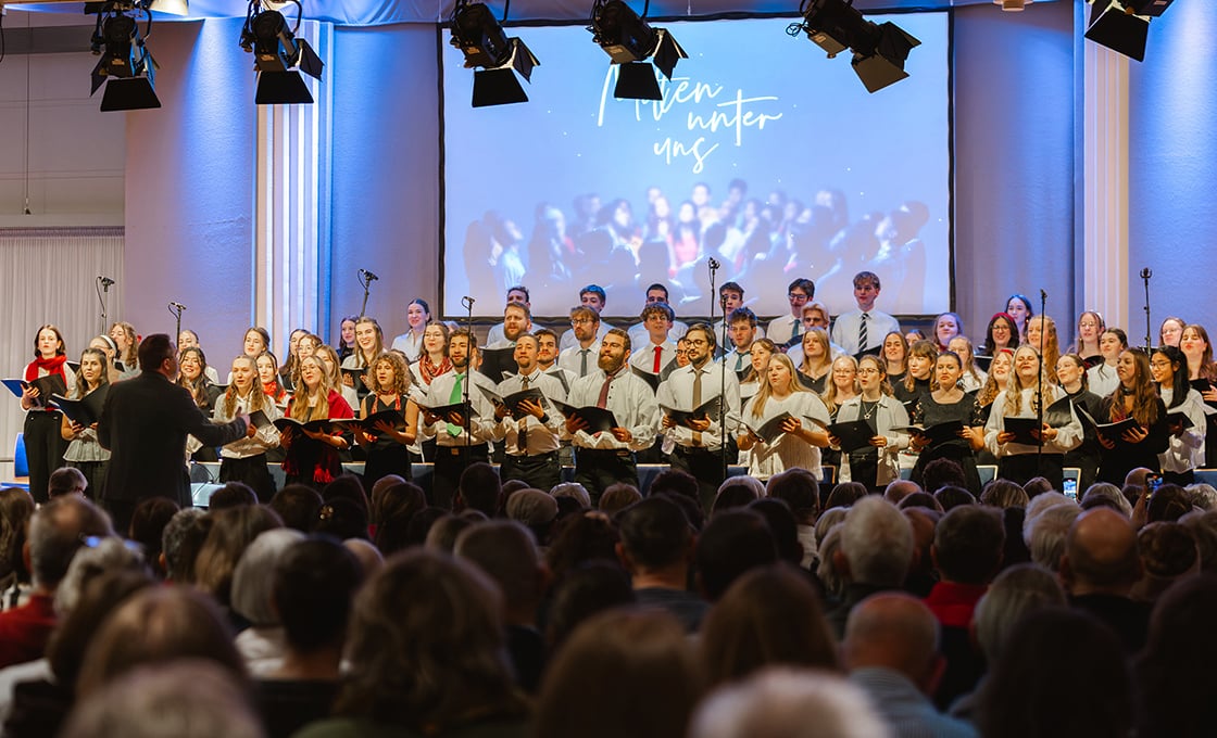Ein Chor tritt auf der Bühne vor einem Publikum auf, mit einem Dirigenten an der Spitze und einer Leinwand, die Heaven Winter Uns zeigt.