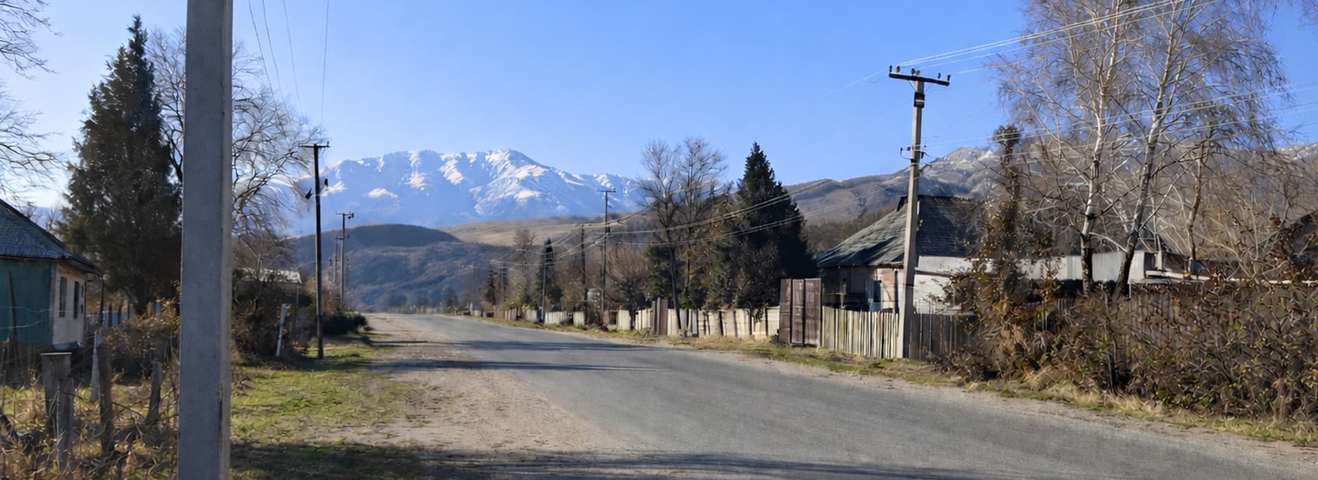 Eine ruhige Landstraße, gesäumt von Häusern und Bäumen, mit schneebedeckten Bergen im Hintergrund unter einem klaren blauen Himmel.